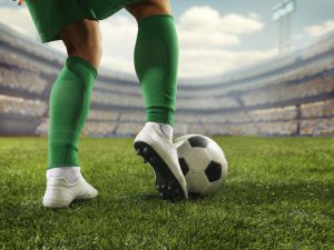 A male soccer player is attempting to kick a ball. The stadium is blurred behind him. The weather is sunny. The sky is cloudy.