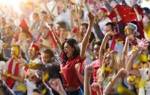 On the foreground a group of cheering fans watch a sport championship on stadium. One girl stands with his hands up to the sky. People are dressed in casual cloth.
